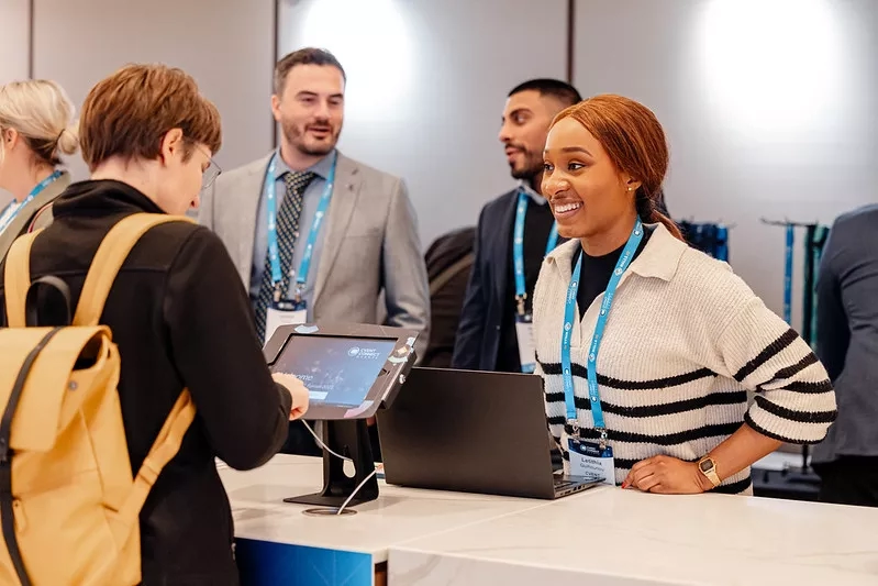 Two people at a booth with a laptop and a tablet, one of them wearing a backpack.