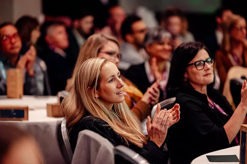 Two women clapping their hands at a meeting in a room with other people sitting at tables.