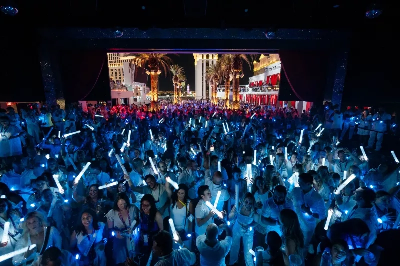 A crowd of people at a night event with a stage and palm trees in the background.