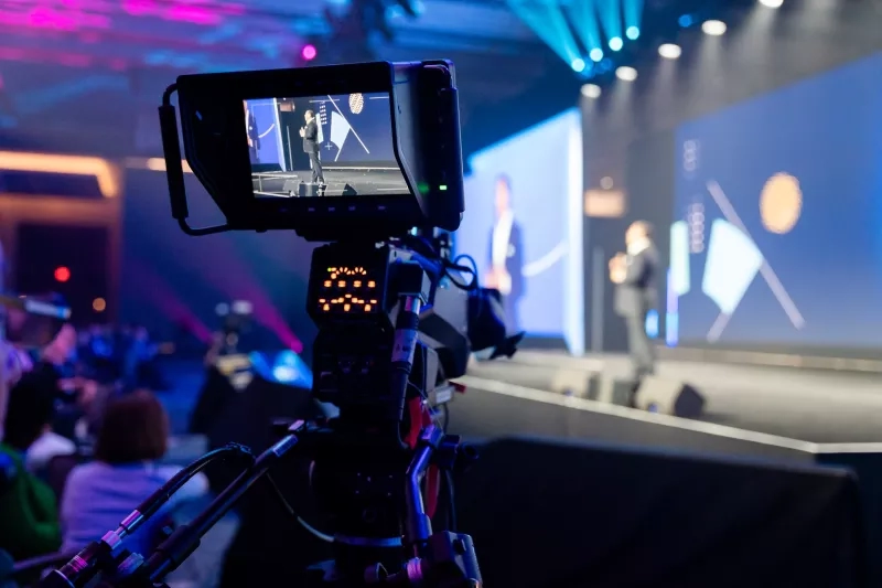 A video camera recording an event with a large audience in front of a stage.