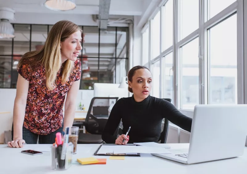 Two women are working on a laptop while conversing in a glass-walled office.