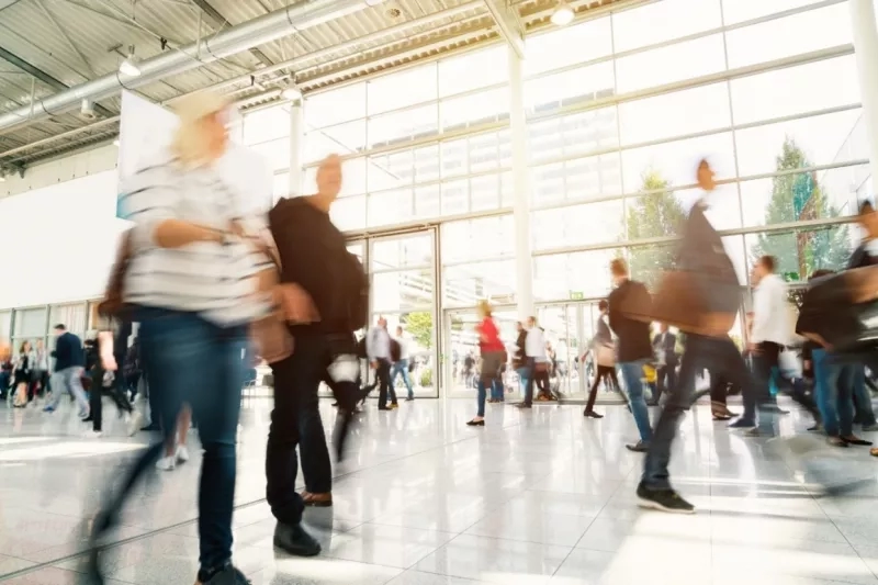 A group of people walking in the blurred corridor of a convention center with glass walls.