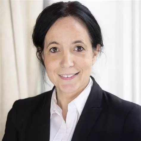 A smiling woman in a black blazer and white shirt stands against a white curtain background.
