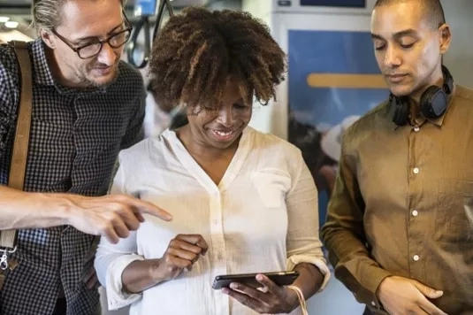 Three people looking at a smartphone, one wearing headphones, in a modern workspace with a poster on the wall.