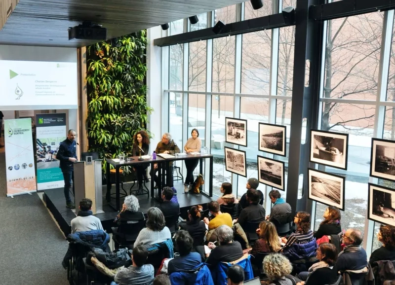 Three speakers sit at a table while a man speaks from a podium in front of an audience.