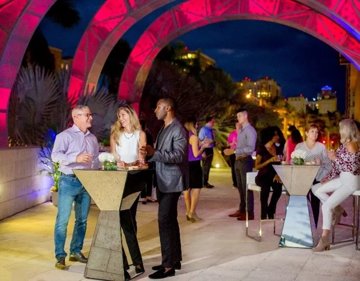People gather at an outdoor event in front of a colorful archway with buildings in the background.