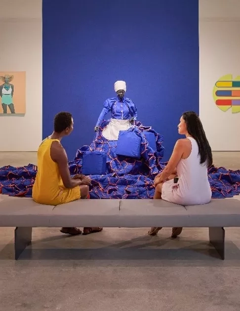 Two people sit in front of a statue on a bench in a gallery space.