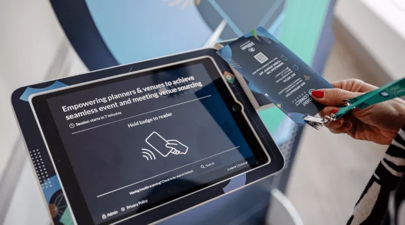 A person holds a badge in front of a tablet screen displaying an event page.