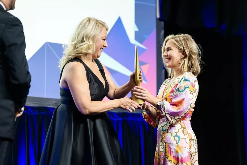 Two women in black and floral dresses hold an award, with a man in a suit nearby.