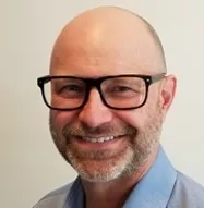 A bald man with a beard wearing black glasses and a blue shirt is smiling at the camera.