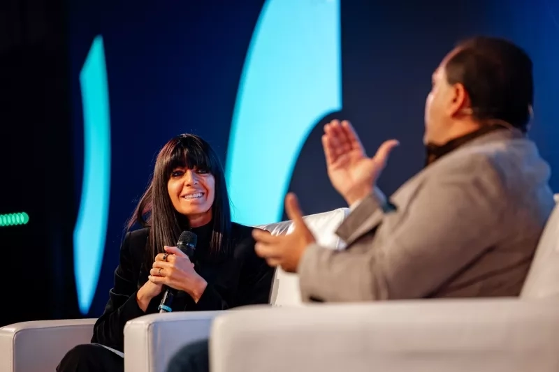 Two people on stage at a conference, one speaking and the other gesturing with his hands.