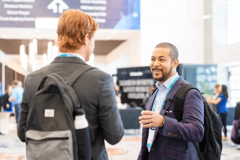 Two men talking in the hallway of a business travel conference with a banner in the background.