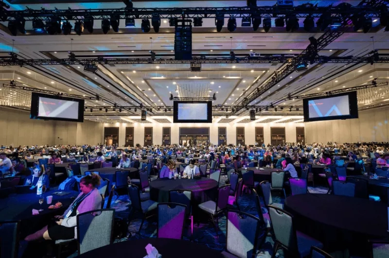 Several people are sitting on chairs around the tables in a huge conference hall with a stage and monitors.