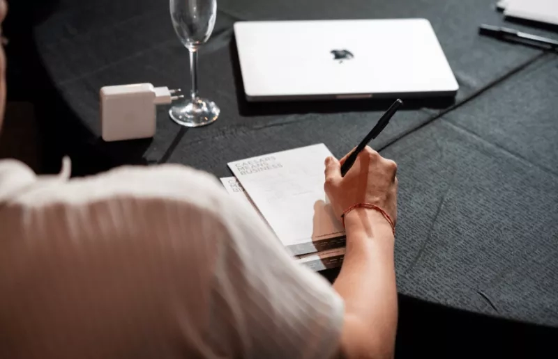 A person with a pen and notebook writing meeting minutes at a round table with a laptop.