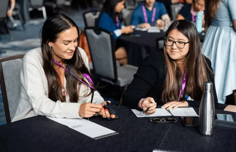 Two women sit at a table during a meeting, discussing and taking notes with a pen and paper.