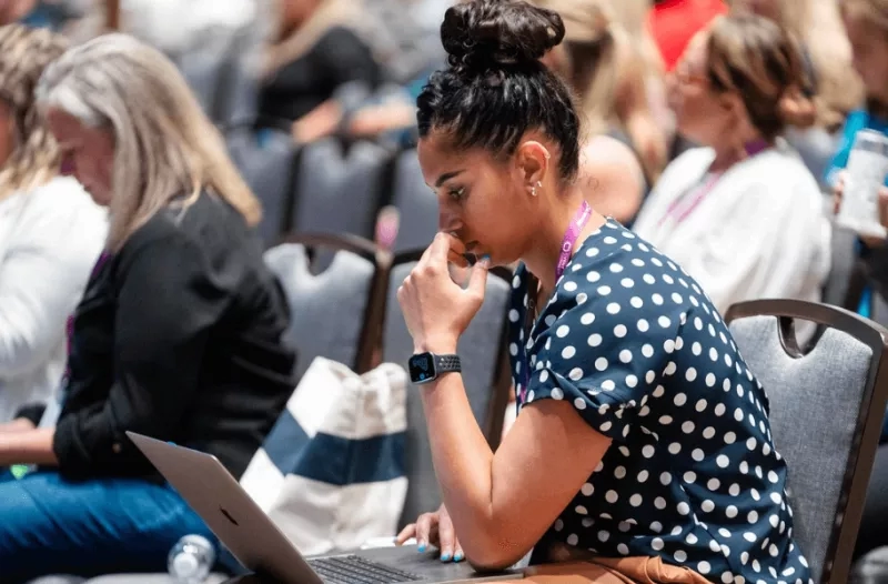 A woman working on her laptop at a conference with other women in the background.