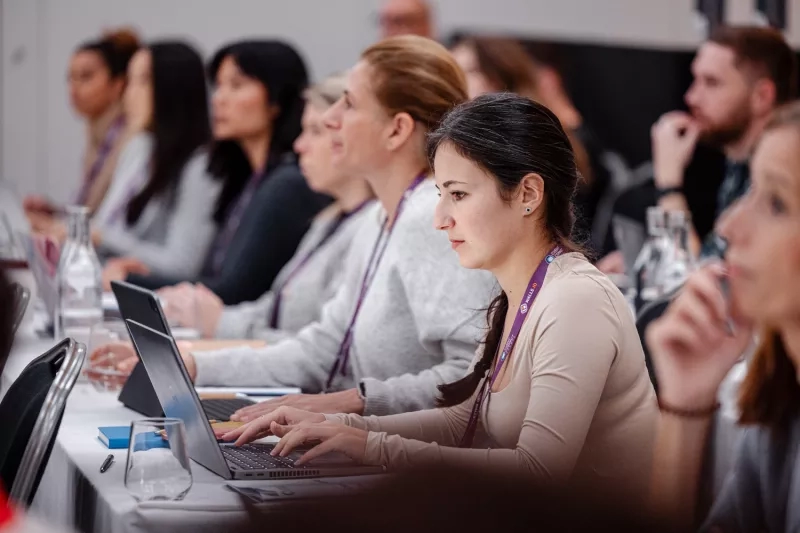 A group of women are sitting at a table and using laptops, with a man behind them.
