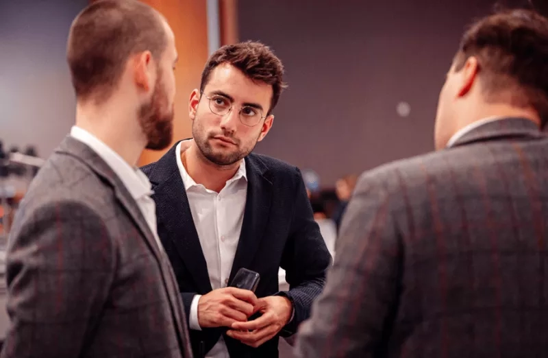 Three men in formal wear are seen in a speed networking event with one man holding a wine glass.