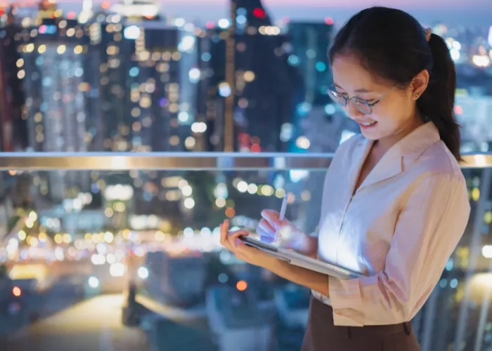 A woman stands on a balcony with a tablet, overlooking a city skyline at night.