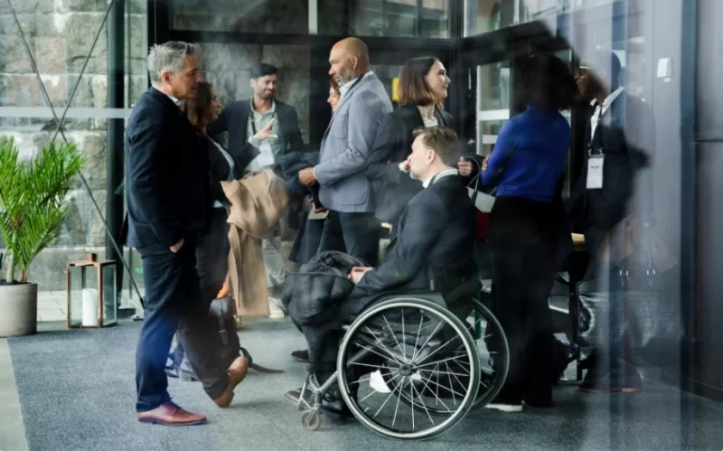 Person in wheelchair is at event, surrounded by others in suits and professional attire, with a stone wall backdrop.