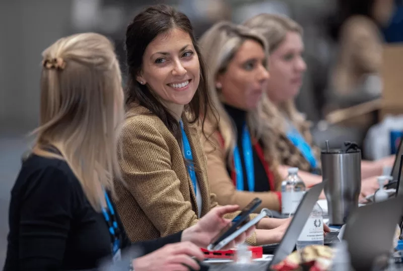 Four women at a conference table with laptops, bottles, and a travel mug in front of them.