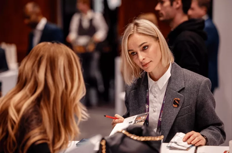 Two women sit at a desk with pamphlets as people mill about in the background.