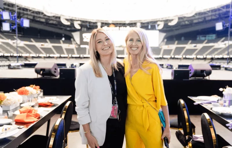 Two women stand together in an empty stadium with dinner tables and chairs set up.