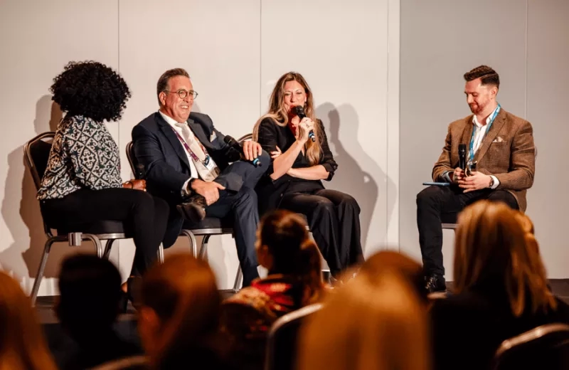 Four people sitting on chairs and talking in a conference room with other people watching them.