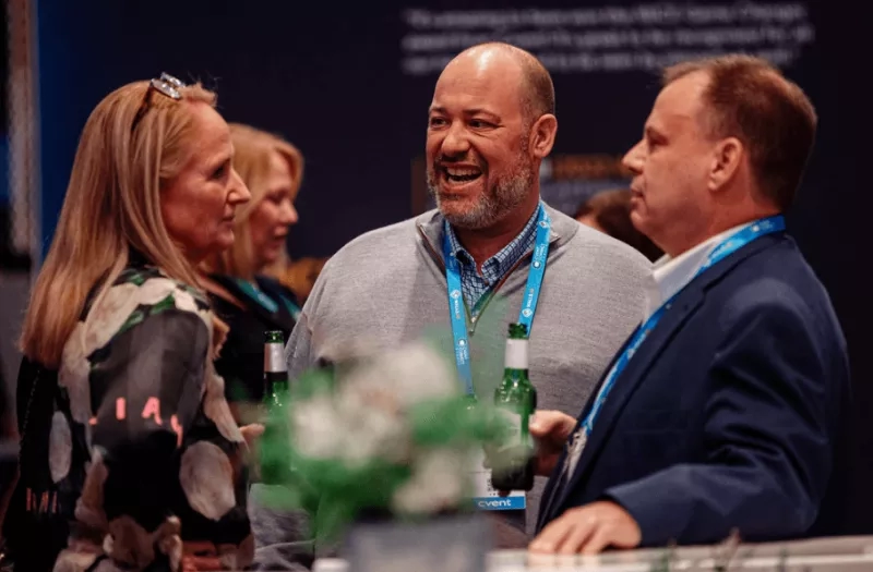 Three people smiling and laughing at an event with two bottles of beer on the table.