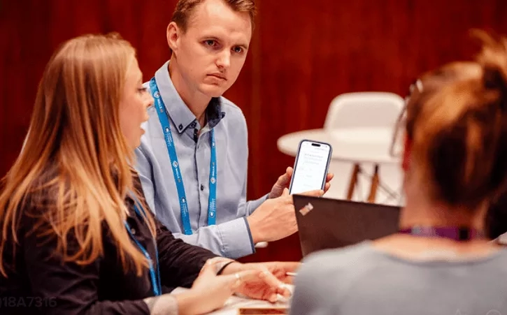 Three people are sitting around a table, one holding a smartphone, and another is typing on a laptop.