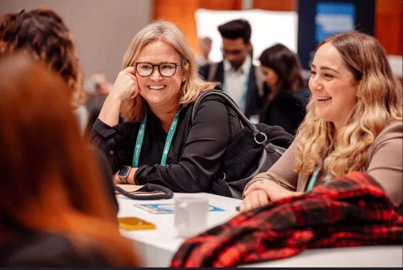 Two women with glasses and a watch are smiling and sitting around a table at a meeting.
