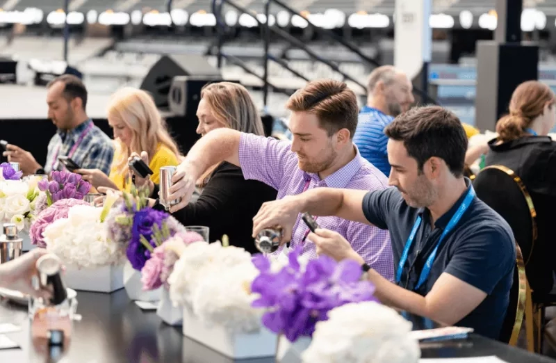 Two men are mixing drinks at an event in an arena with purple and white flowers.