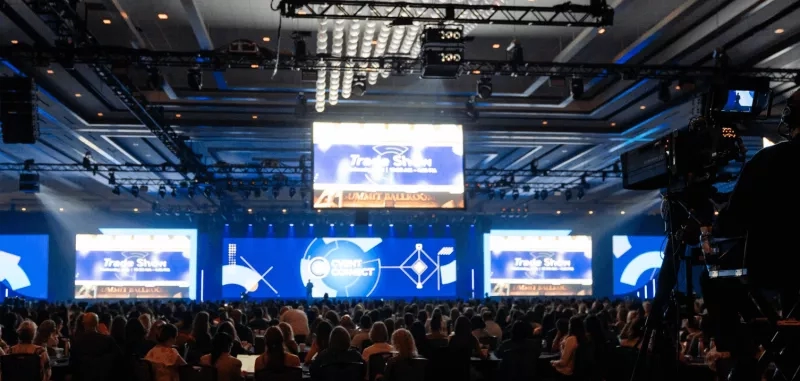 Trade show conference theme with a large crowd and a stage with several screens and speakers.