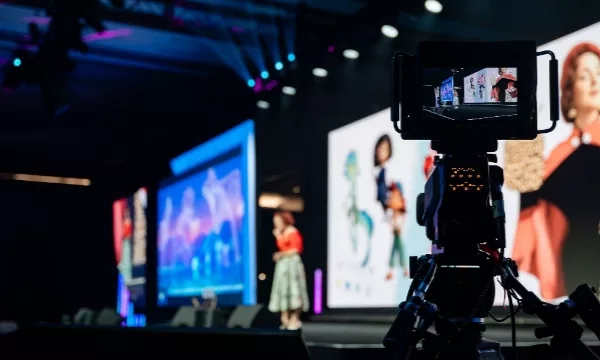 Video camera recording a woman standing on stage with a spotlight on her.