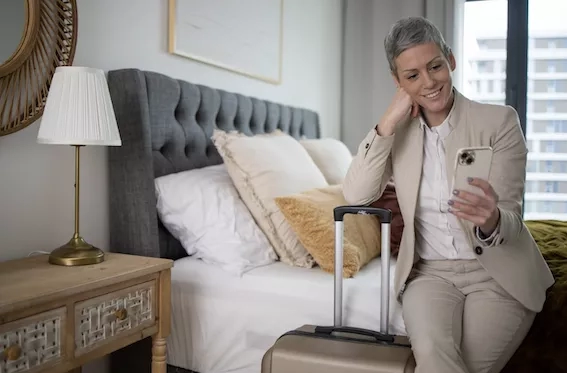 A woman with grey hair is sitting on a bed using her phone in an apartment.