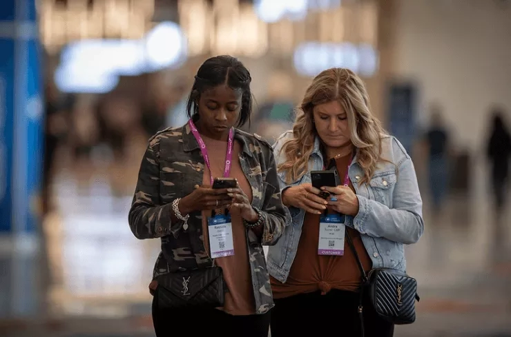 Two women in a hall with cell phones and name tags around their necks, looking down.