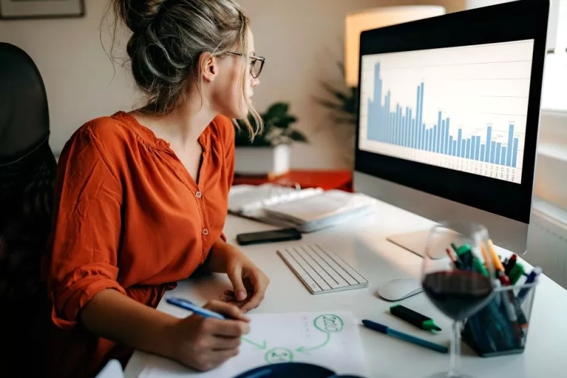 A woman is sitting at a desk with a computer and wine, working on a business plan.