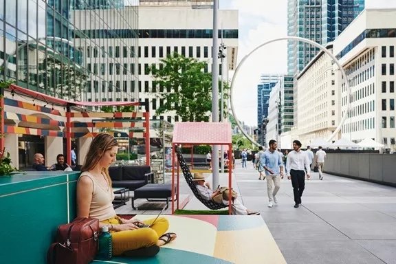 A woman is sitting on a bench in the outdoor area with people walking by, and buildings are behind her.