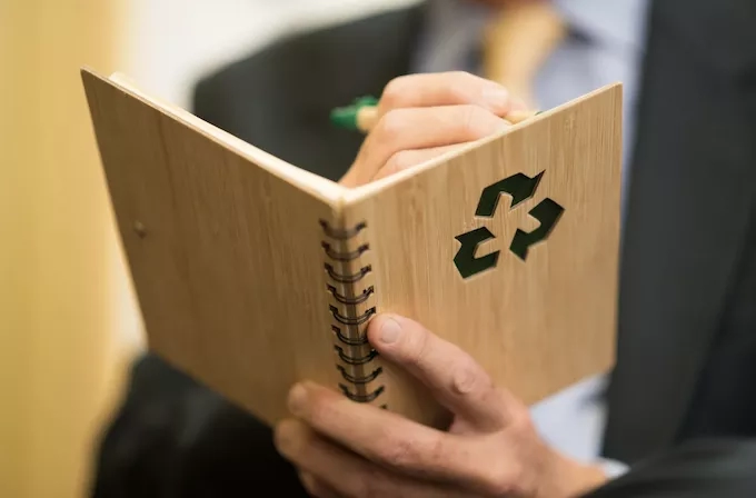 A person wearing a business suit holds a spiral notebook and writes with a green pen.