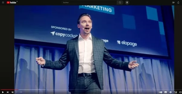 A man in a suit giving a speech in front of a large blue screen.