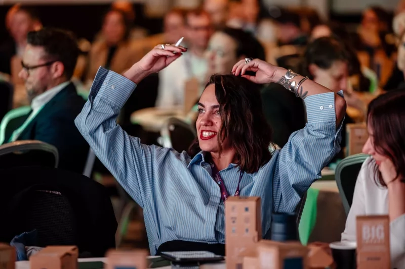Woman sitting at a table raising her arms above her head and smiling at a conference.