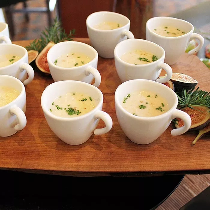 Several white mugs with cream soup, garnished with herbs, on a wooden board, with a blurred background.