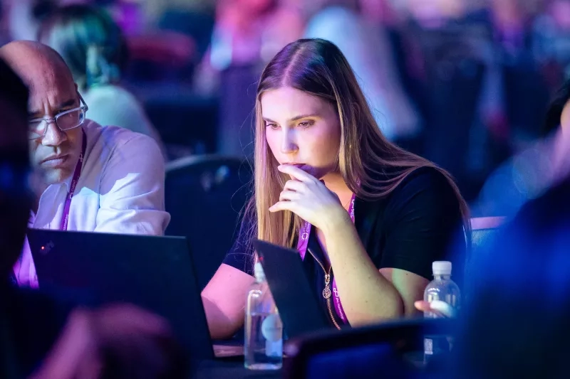 A woman with a laptop looks pensively at a large event with other people in the background.