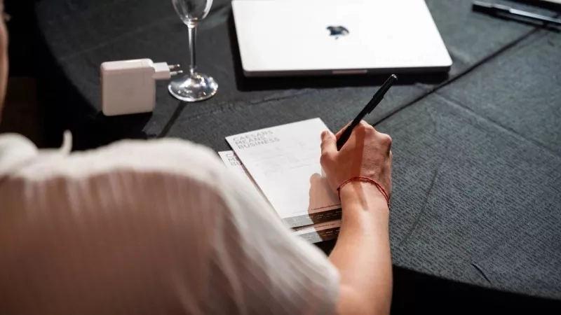 A man signing a paper at a table with a laptop and a glass of wine.