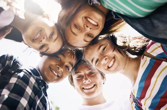 Five young people stand close together, smiling and looking up for the camera, in an outdoor setting.