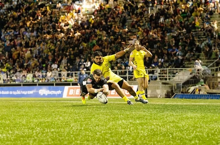 A rugby player is being tackled by two other players during a rugby game in front of an audience.