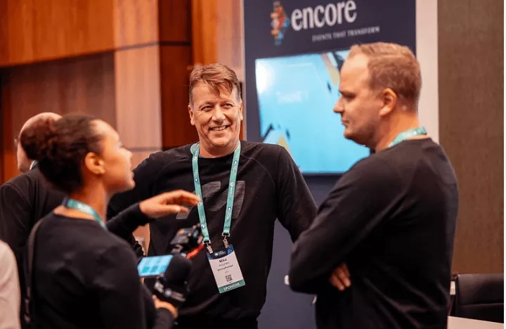 Three people at a conference with a large "Encore" sign in the background, and a table and chairs nearby.
