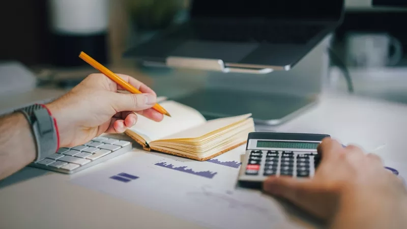 Person writing notes on a notepad while holding a calculator in front of a laptop.