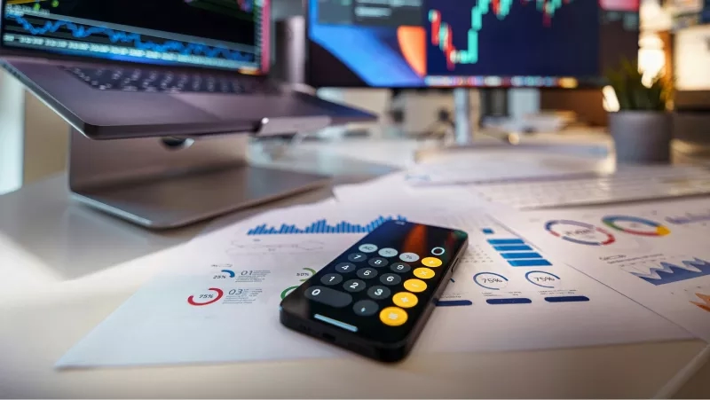 A desk with a remote, documents, a laptop, and a computer with a stock chart on the screen.
