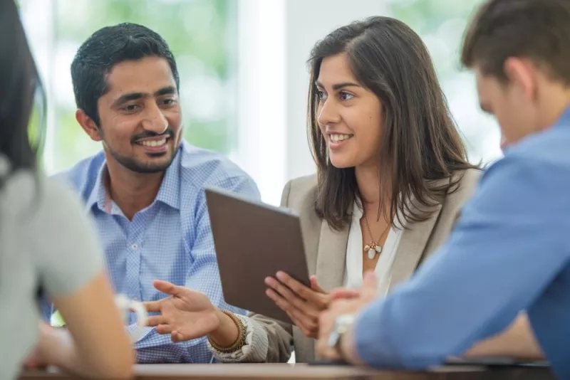 Four colleagues are sitting around a table, one holding a tablet and talking about a business meeting.